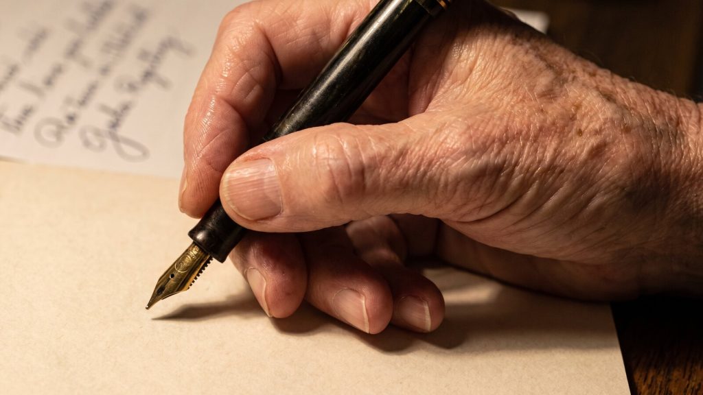 An older person's hand writing in cursive on cream paper with a fountain pen