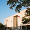 A limestone county courthouse in downtown Austin framed by live oak trees with Texas and U.S. flags