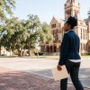A woman carrying a folder walks along a brick sidewalk toward a historic stone courthouse under live oaks in Victoria County, Texas